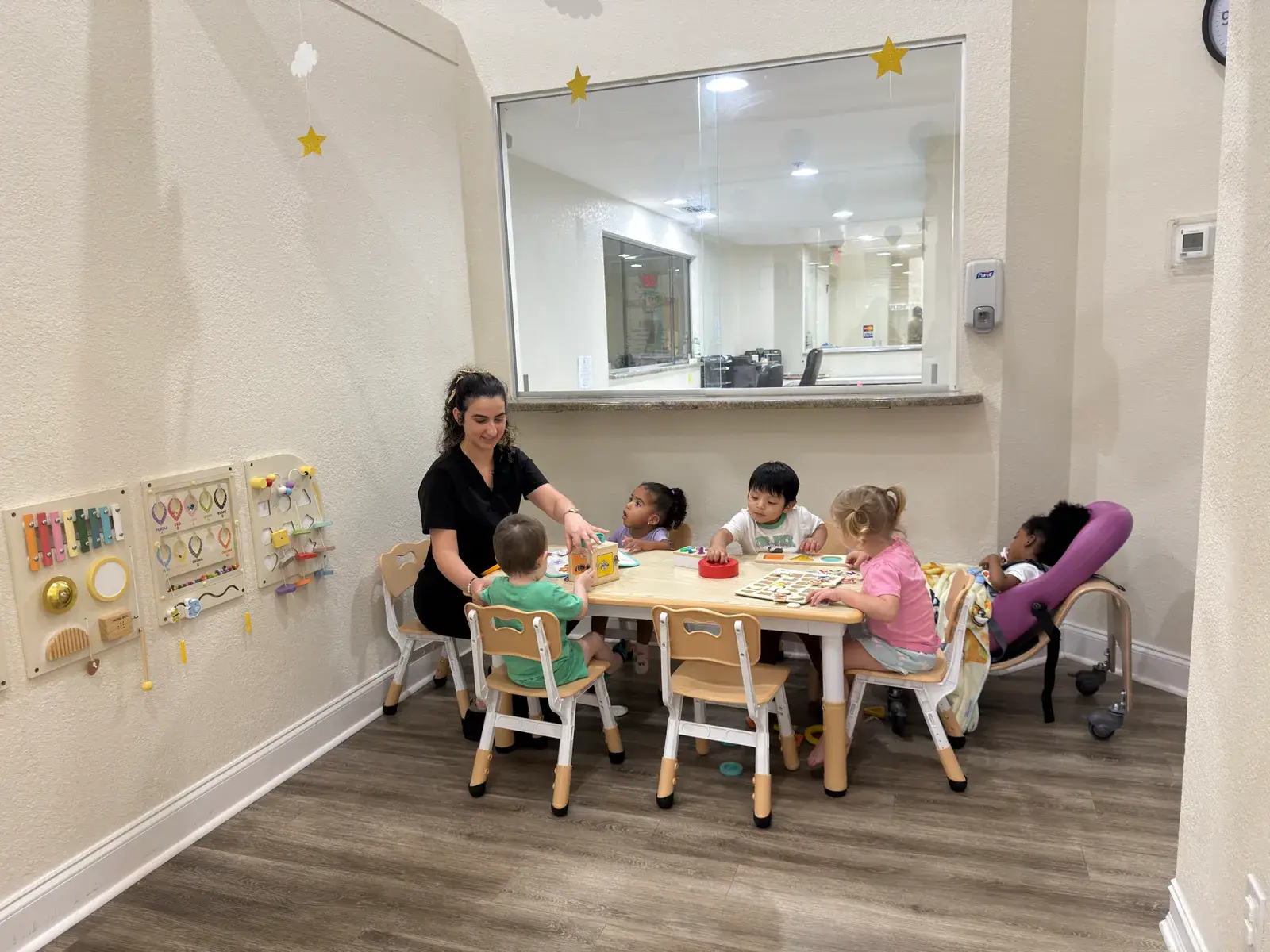 A staff member leading a small group of children at the learning table, with the nurses' station window behind at Ellie's World PPEC in Port Charlotte, Florida